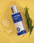 A bottle of bye bye stye face wash sits on a yellow background with a sprig of rosemary and gentle face wash suds.